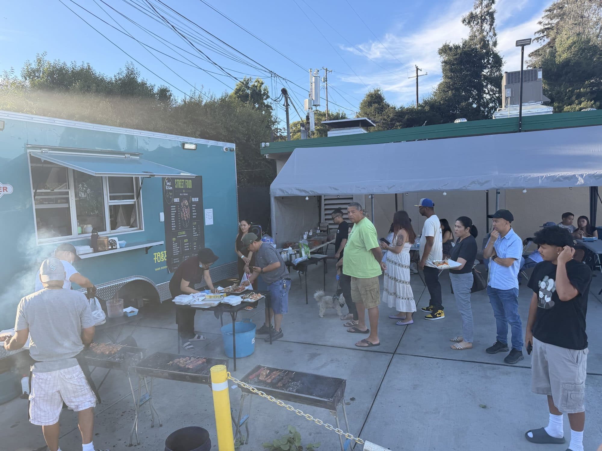 The Tino's Kitchen trailer with the grills going and a line of customers under the tent